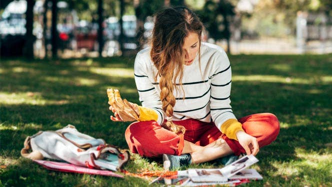 College-age girl sitting on the grass eating a sandwich and reading textbooks