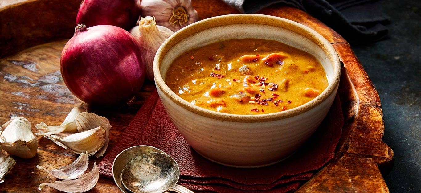 Shiro Wat (Chickpea Flour Stew) in a white ceramic bowl surrounded by red onions and garlic on a wood tabletop