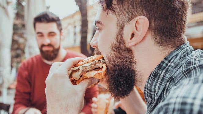 Young man eats a cheeseburger while another man in the background watches him