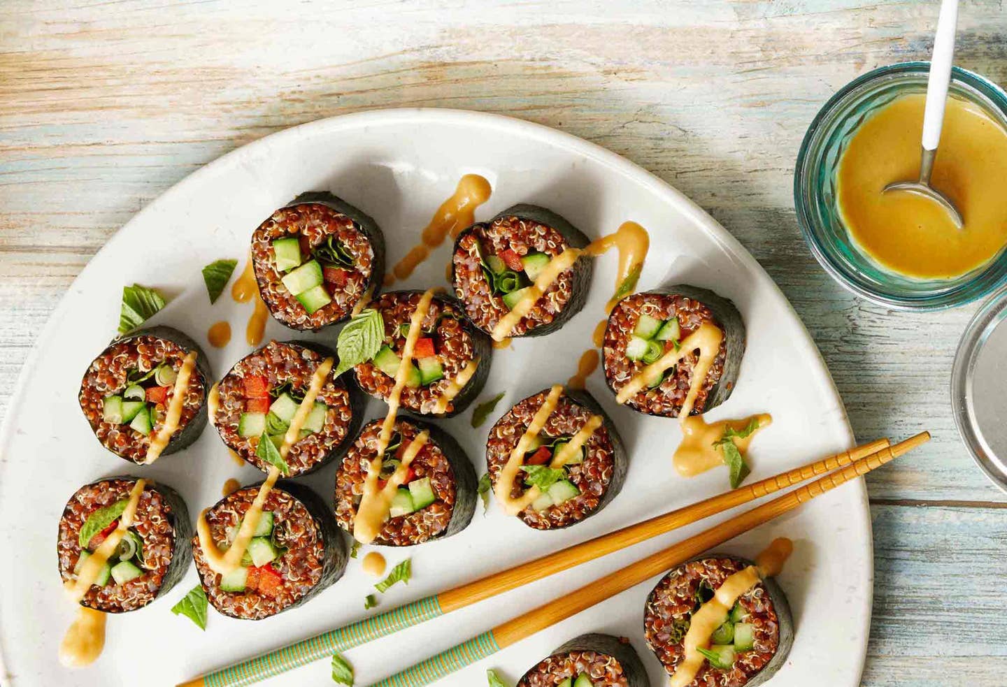 A platter of vegan sushi rolls filled with quinoa, carrots, and cucumber, drizzled with a Thai peanut sauce, with additional sauce in a bowl to the side