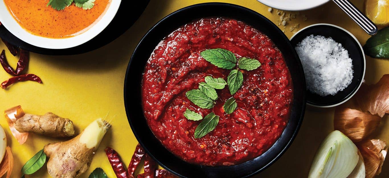 Homemade Harissa in a dark bowl against a yellow background surrounded by fresh ginger, chiles, and onion slices