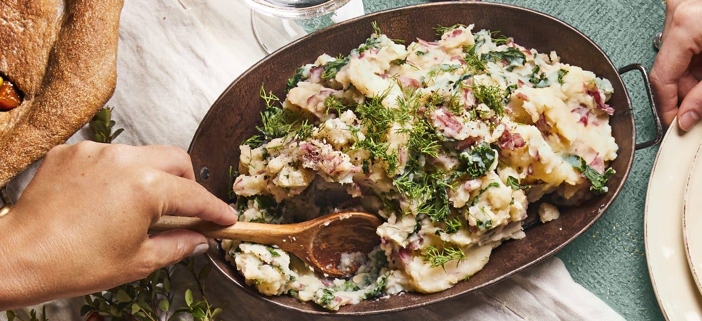 Mashed Potatoes with Kale and fresh dill in a metal serving dish being stirred by a woman's hand