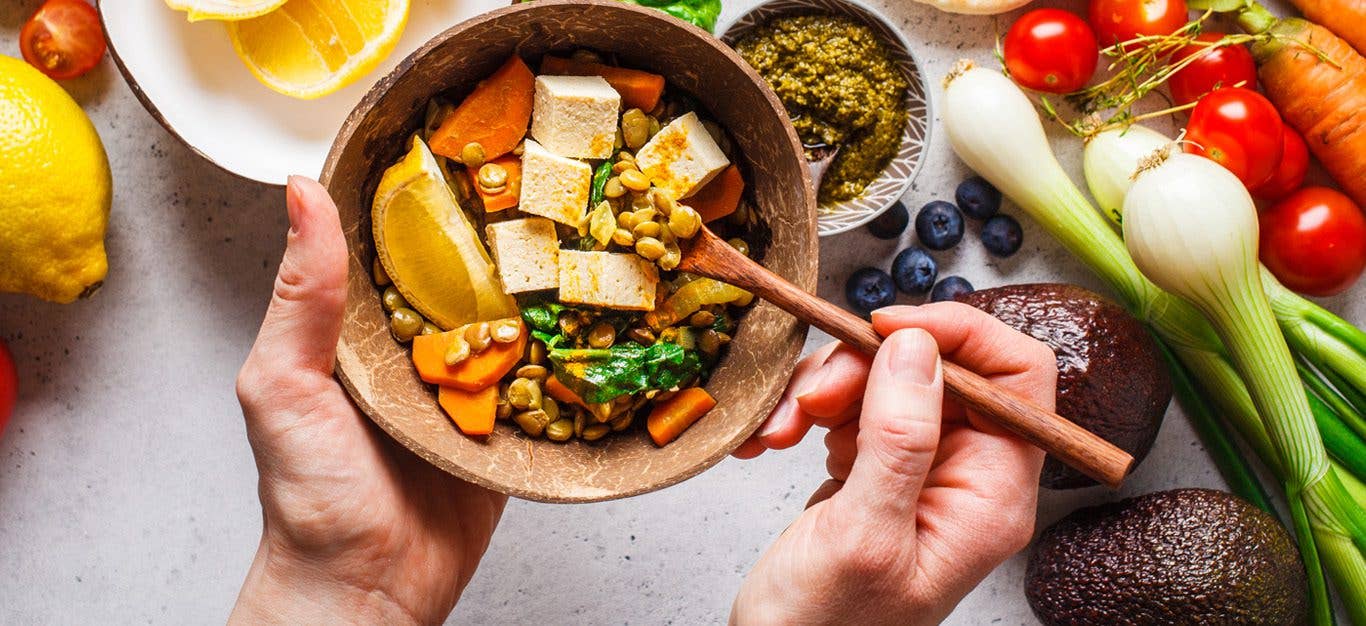 Man holds up bowl with colorful array of vegetables, tofu, grains, and lemon wedge, while more fruits and veggies are on the table below