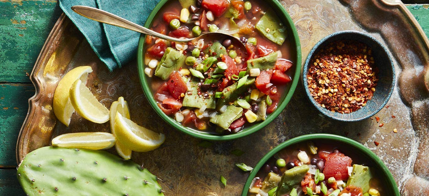 Sopa de Nopales in green bowls surrounded by lemon wedges, cactus paddles, and red pepper flakes