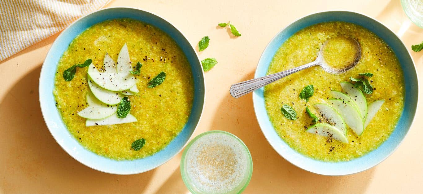 Sunshine Gazpacho topepd with apple slices and mint in blue bowls on a peach colored tabletop