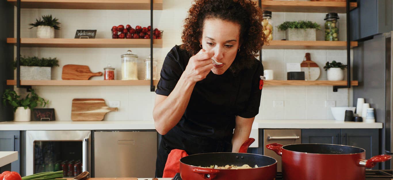 a woman stands over a sauté pan on her stove, with a wooden spoon to her mouth, tasting what she's cooking