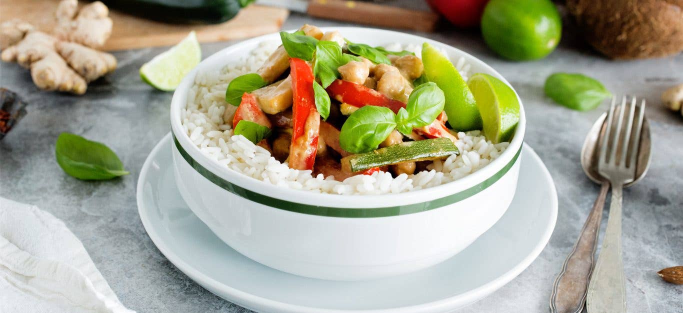 Thai Chickpea-Almond Curry in a white bowl surrounded by fresh limes wedges, basil, and ginger root