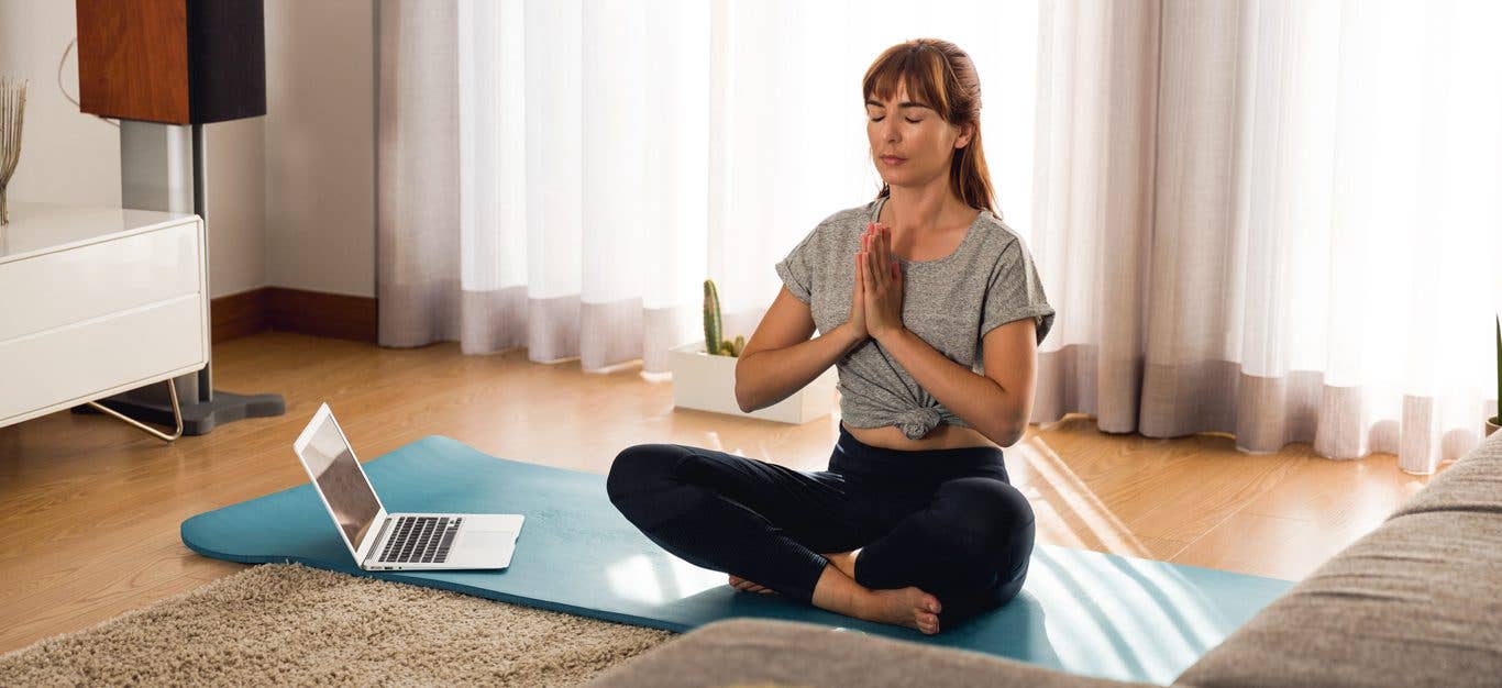 Woman sitting on yoga mat in her home meditating with an open laptop on the floor